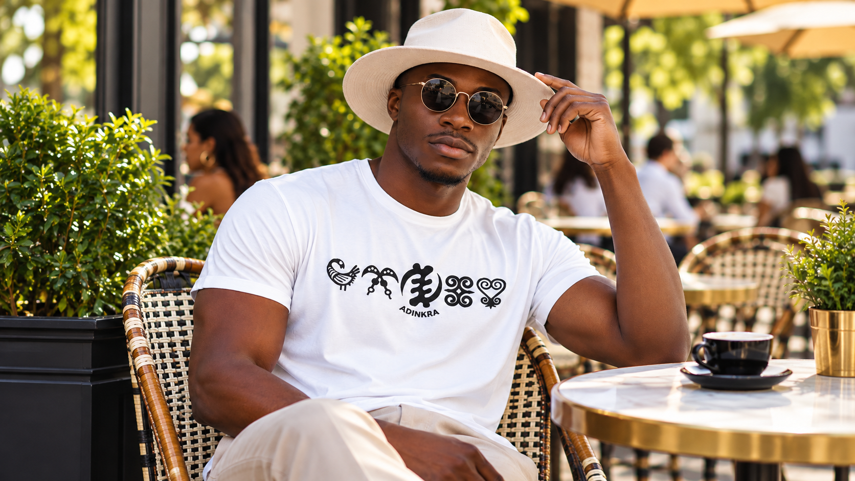 Model wearing a white Adinkra T-shirt and hat at an outdoor café